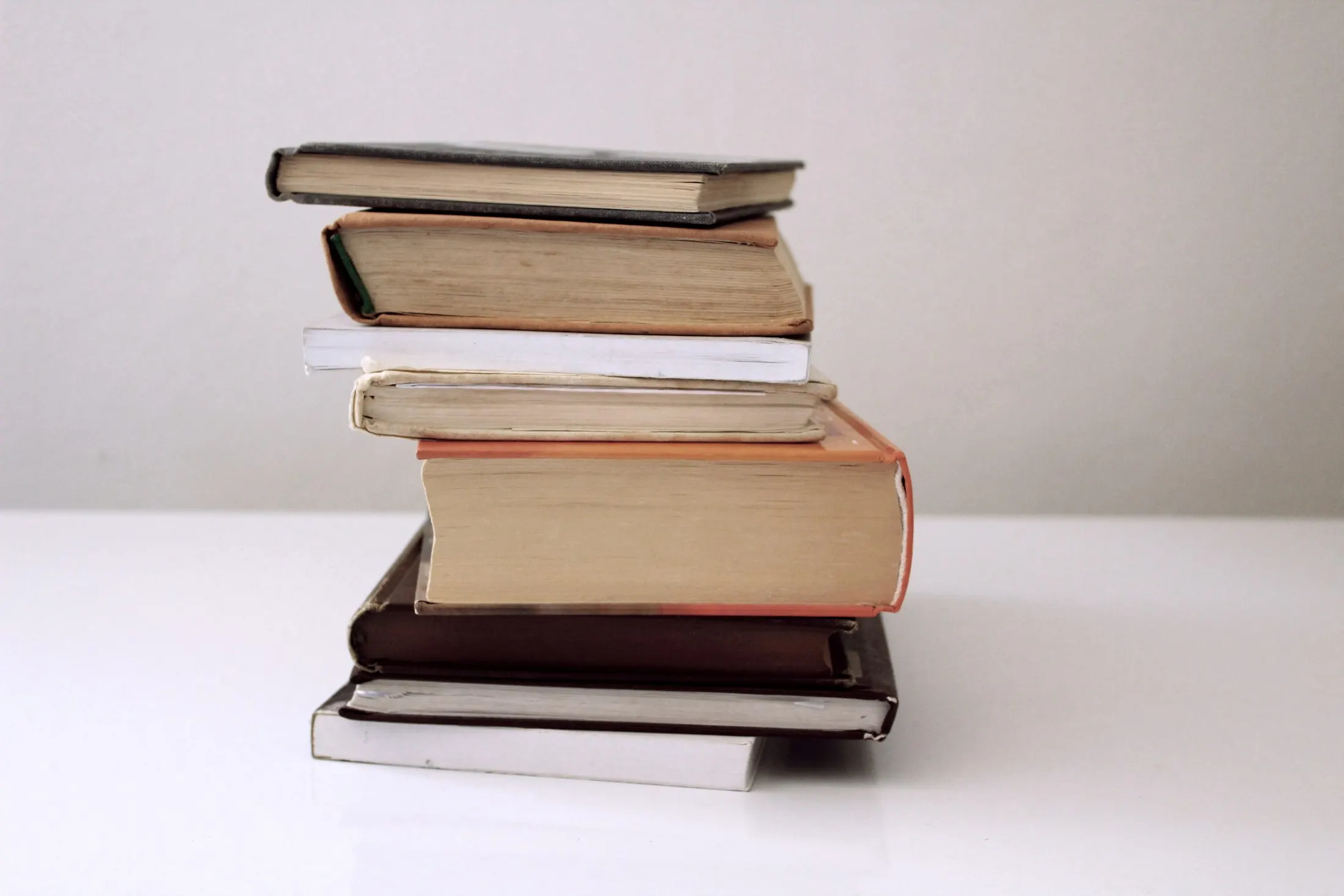 A loose stack of eight worn hardback and paperback books, with no titles visible, on a white tabletop against a plain pale-grey wall.