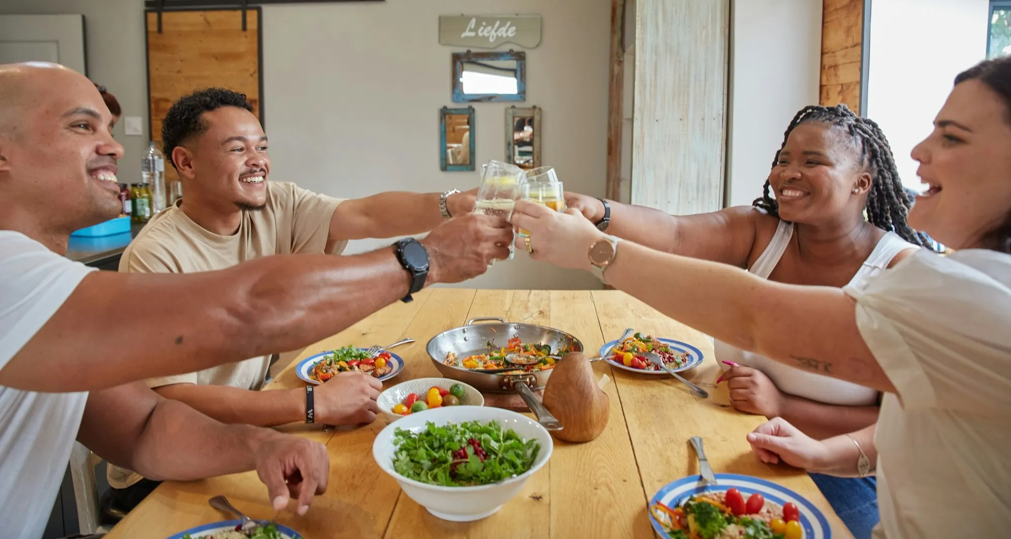 Friends raising their glasses together over a long wooden table of shared dishes in a bright, modern home.