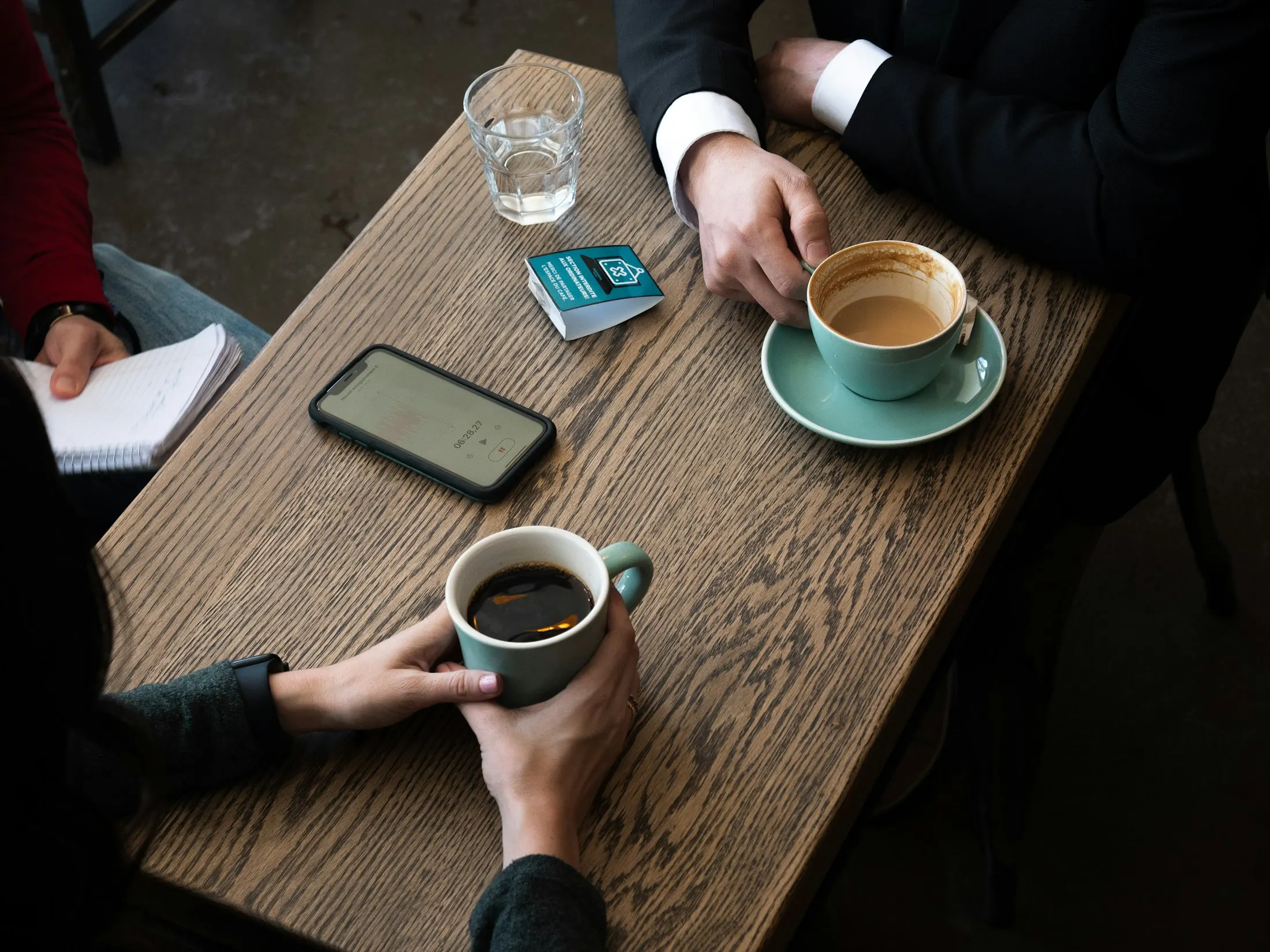 Overhead view of a wooden café table during a conversation: one person holds a green mug of black coffee, another in a dark suit rests a hand beside a latte on a saucer, a phone shows a voice-recording screen, and a third person at the left edge holds a notebook.
