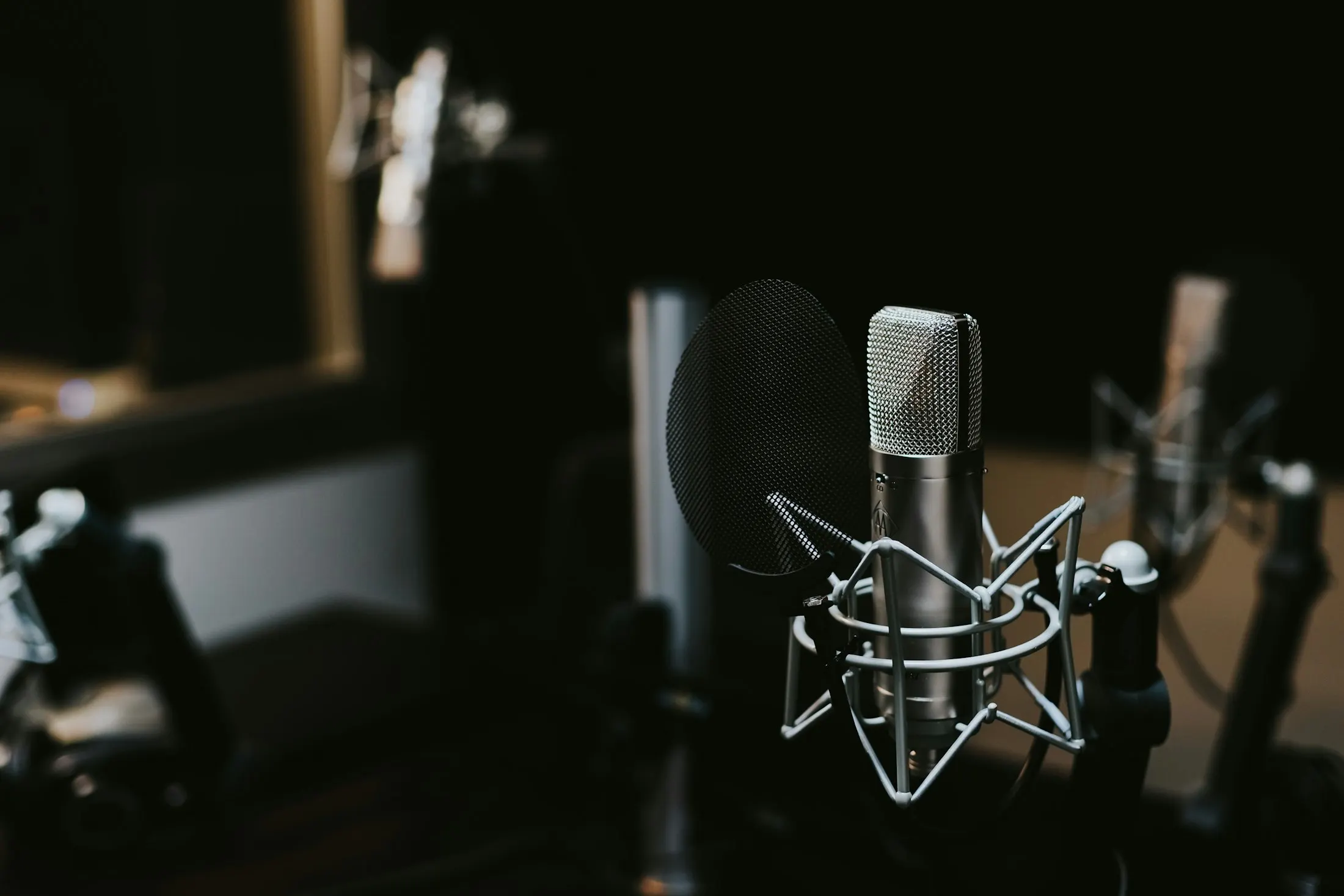 A silver condenser microphone in a shock mount with a pop filter in a dim recording studio, with more microphones softly out of focus behind.