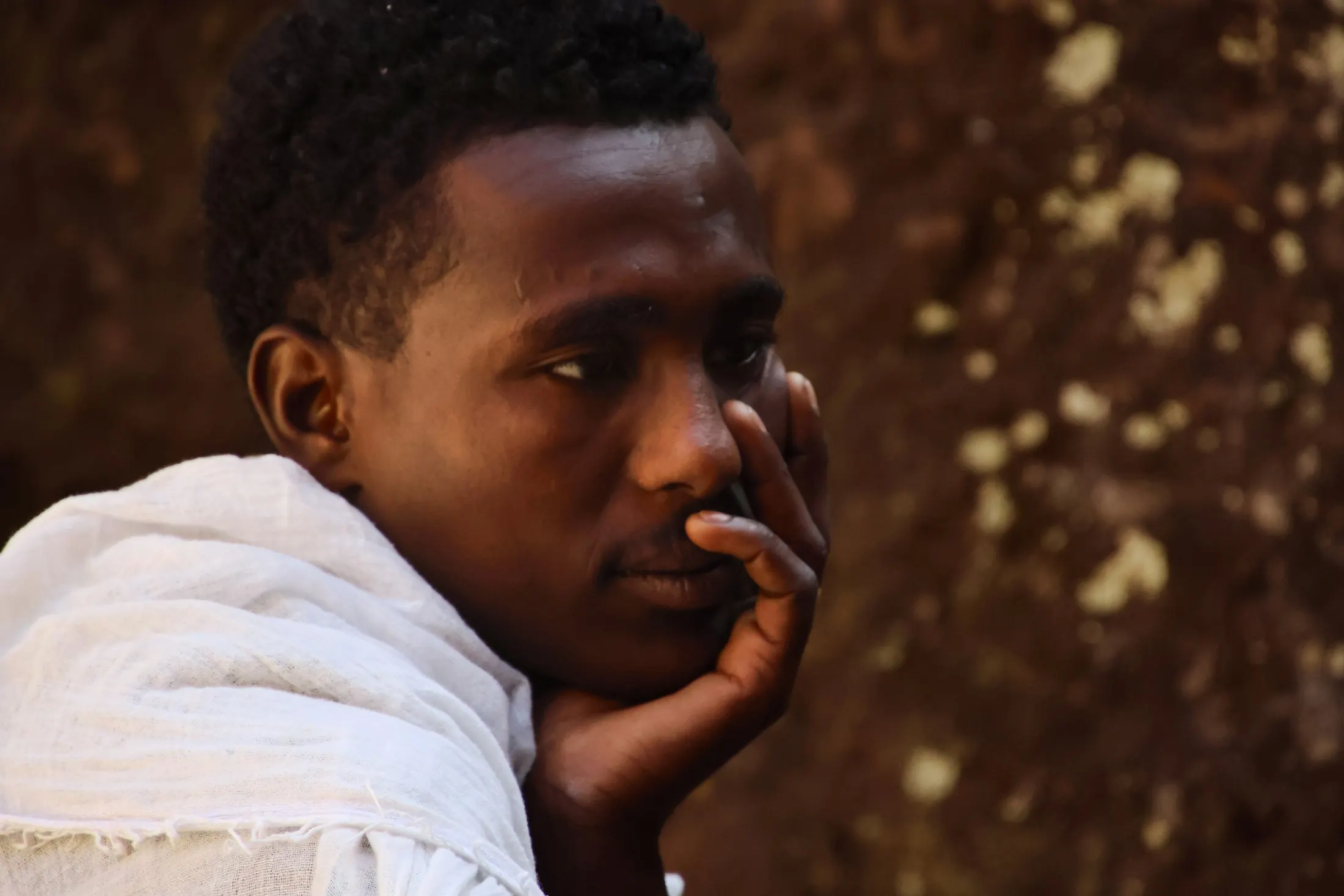 Candid portrait of a young man with a white cotton shawl over his shoulder, resting his cheek on his hand and looking thoughtfully off to the side in front of a textured, mottled brown wall.