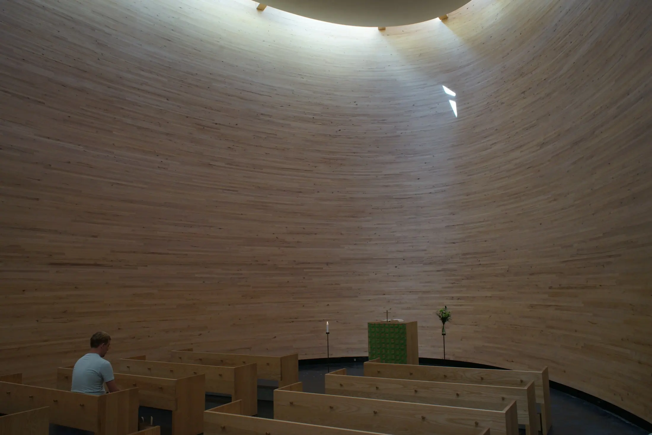 A quiet timber chapel lit softly from a skylight, with one person seated alone in the pews