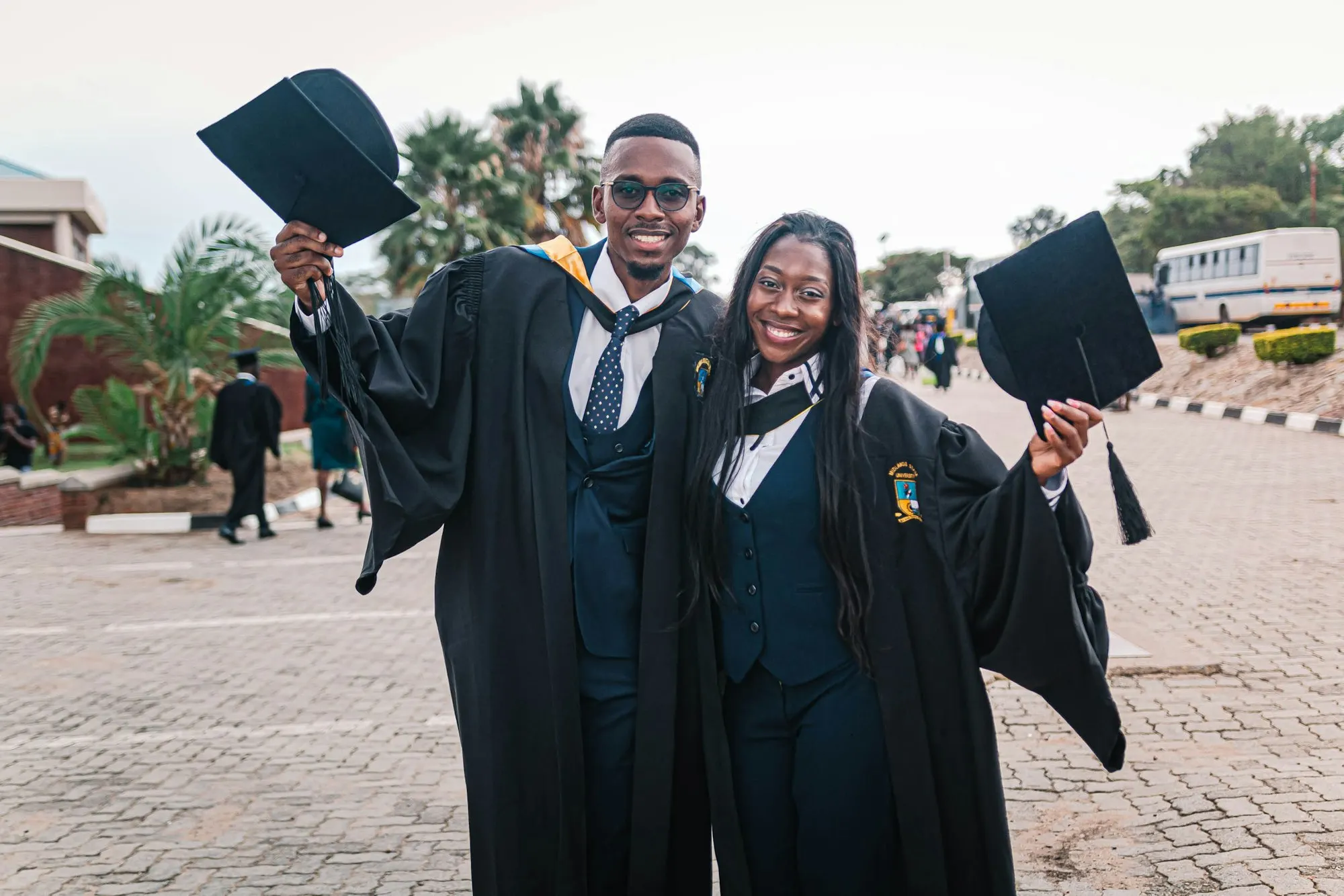 Two smiling graduates in academic gowns raising their caps on a sunny campus.